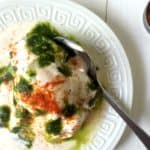 A white plate with dahi vada, also known as dahi bhalla, topped with yogurt, green chutney, red chili powder, and fresh cilantro, with a spoon resting on the side. The plate is on a white wooden surface.