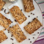 Crunchy Nut Free Granola Bars with oats and chocolate chips are arranged on a white plate. A metal measuring cup filled with oats and scattered chocolate chips add to the scene. The plate sits atop a striped, colorful cloth.
