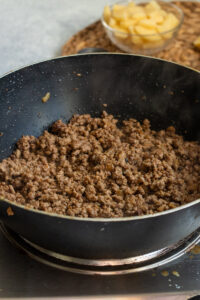A black frying pan on a stovetop filled with cooked Qeema, steam rising; in the background, a glass bowl contains chopped potatoes on a woven mat, hinting at an Aloo Keema recipe in progress.