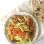 A white plate with stir-fried Aaloo Gajar—spicy carrots and potatoes—garnished with black sesame seeds, sits next to folded pieces of flatbread on a white napkin atop a light-colored table.