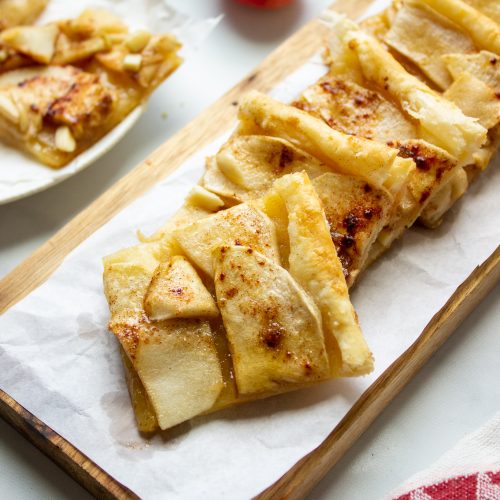 Slices of puff pastry apple tart with a golden, flaky crust are arranged on a wooden board lined with parchment paper. A red apple and a red-and-white cloth are visible in the background.