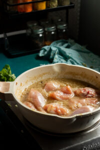 Raw chicken pieces simmer in a white skillet with a bubbling sauce, hinting at a delicious white chicken curry; spice jars and a teal kitchen towel add color to the background.