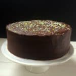 A One Bowl Chocoholics Chocolate Cake with smooth frosting and colorful sprinkles on top, displayed on a white cake stand against a black background.