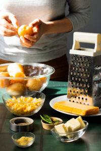 A person peels a potato over a bowl of whole potatoes, prepping for Aloo Cutlets. Nearby are bowls of grated cheese, butter, spices, green chilies, and a cheese grater on a green countertop in bright sunlight.