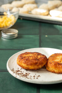 Two golden-brown Aloo Cutlets sit on a white plate, with some crumbs nearby. In the background, there is a glass bowl of shredded cheese and a baking tray with more Potato Cutlets Recipe items, slightly out of focus.