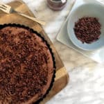 A decadent chocolate mousse tart topped with chocolate shavings sits on a wooden board next to a small bowl of extra shavings and a jar of chocolate sauce on a marble surface. A fork rests beside the tart.