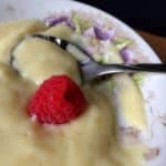 Close-up of a spoonful of creamy custard on a floral-patterned plate, with a single fresh raspberry placed on top for an elegant touch.