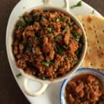 A bowl of spiced chicken karahi qeema garnished with cilantro, served with flatbread and a side of mixed vegetable pickle on a white plate.
