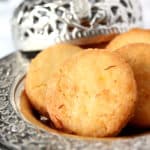 Three round, golden brown meethi tikiyaan are arranged on an ornate silver plate, with an intricately designed silver container in the background. The cookies have a slightly crumbly texture and visible saffron strands.