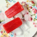 Two red popsicles infused with rooh afza rest on ice cubes atop a white plate with a colorful floral pattern, all placed on a light wooden surface.