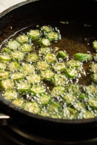 Sliced okra pieces frying in hot oil in a black skillet, with bubbles forming around the okra as it cooks-a delicious start to any Bhindi Masala Recipe.