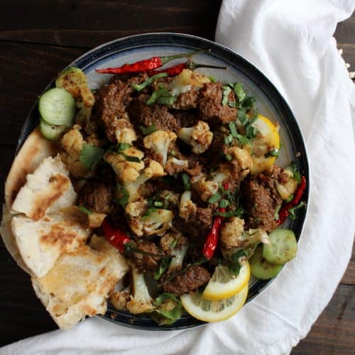 A plate of Gobi Gosht with spiced meat, roasted cauliflower, red chilies, fresh herbs, lemon slices, cucumber, and pieces of flatbread on a dark wooden surface with a white cloth beside it.