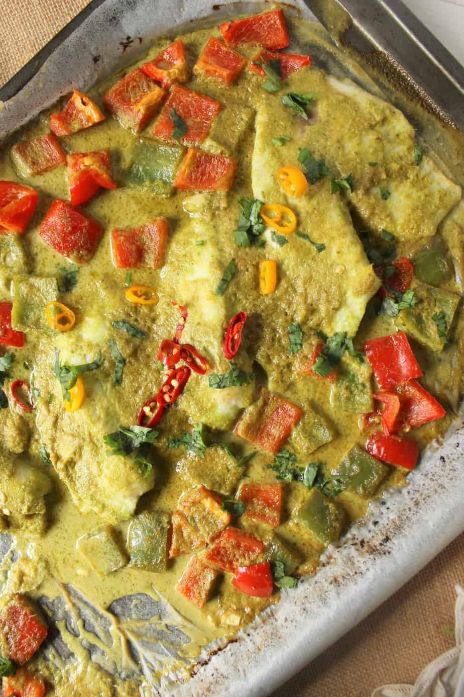 Fish Tray Bake featuring baked fish fillets smothered in green curry sauce, garnished with chopped cilantro, sliced red and yellow chili peppers, and diced red bell peppers on a metal baking tray.