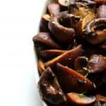 A close-up of a white dish filled with delicious roasted mushrooms, garnished with small bits of green herbs, on a white background.