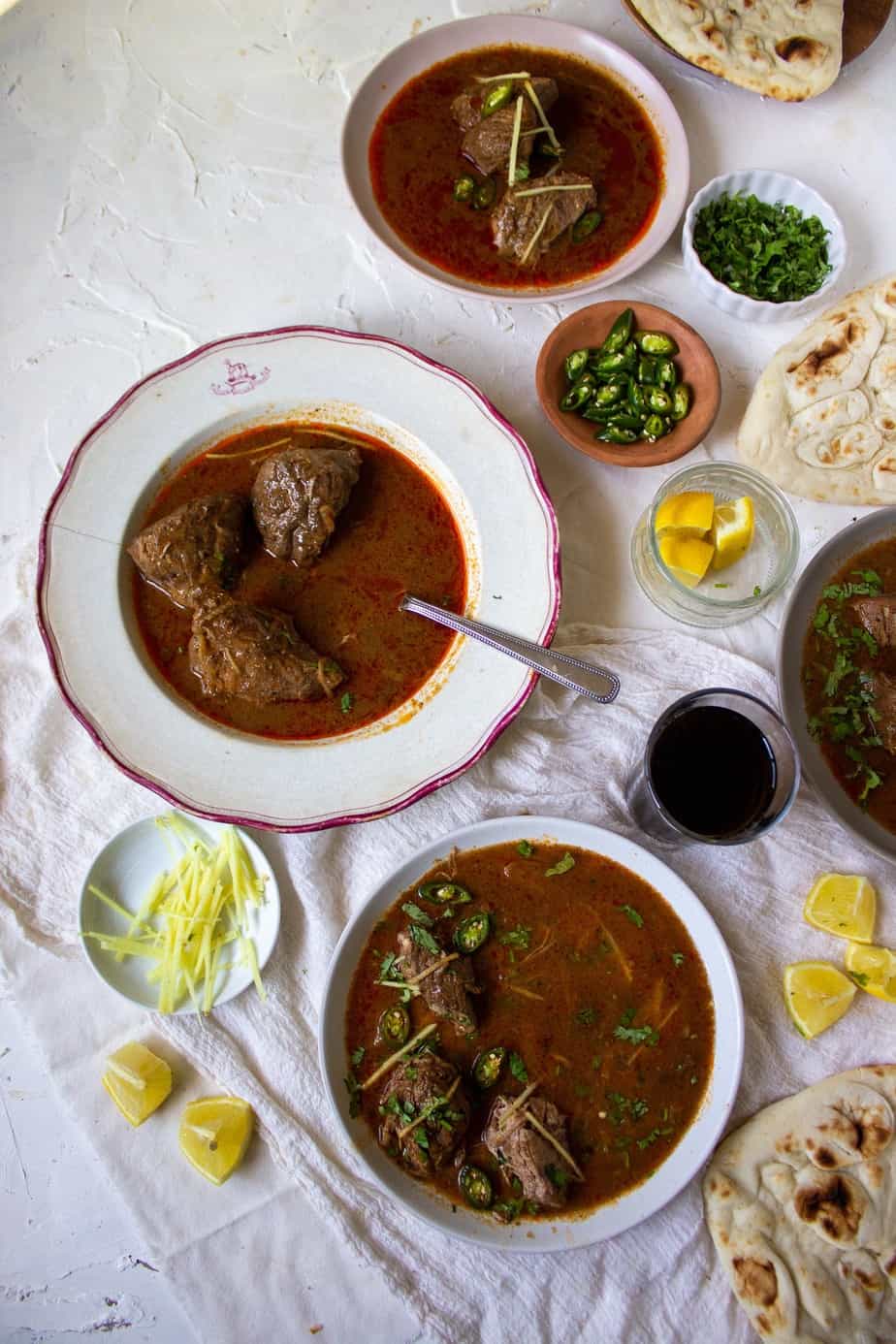 three dishes with nihari in them and little bowls of garnishes and some naan