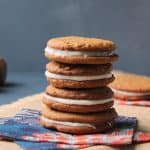 A stack of five sandwich cookies with cream filling sits on colorful napkins atop a burlap cloth, evoking the cozy appeal of Chewy Ginger Cookies, with a blurred cookie and neutral background behind.