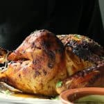 A person in a dark shirt is holding a platter with a large Desi Roast Turkey, garnished with herbs. In the foreground, there is a brown bowl with green sauce.