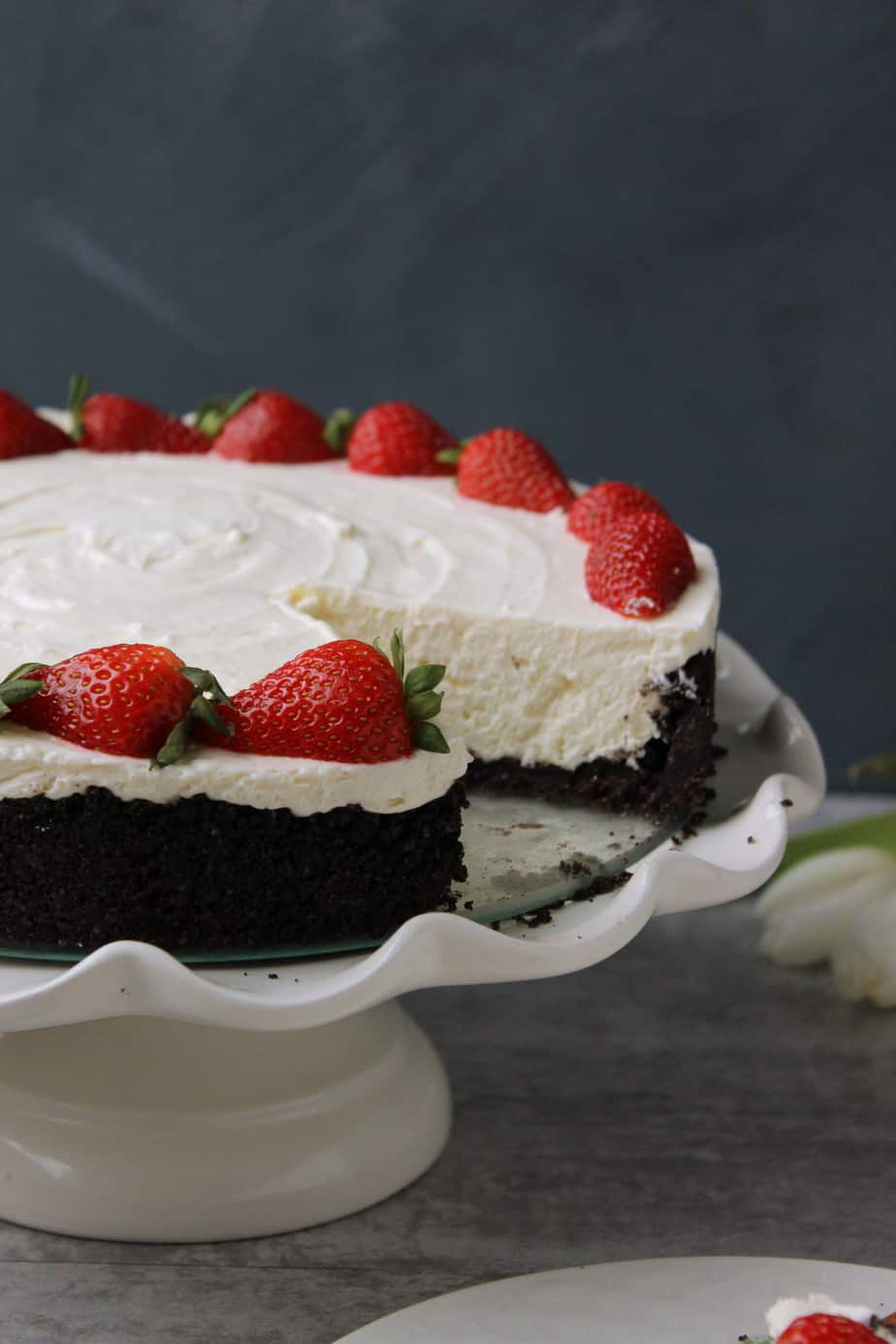 A creamy Best No Bake Oreo Cheesecake with a dark chocolate crust, topped with whole strawberries and displayed on a white cake stand. A slice has been removed, revealing the smooth, rich texture inside.