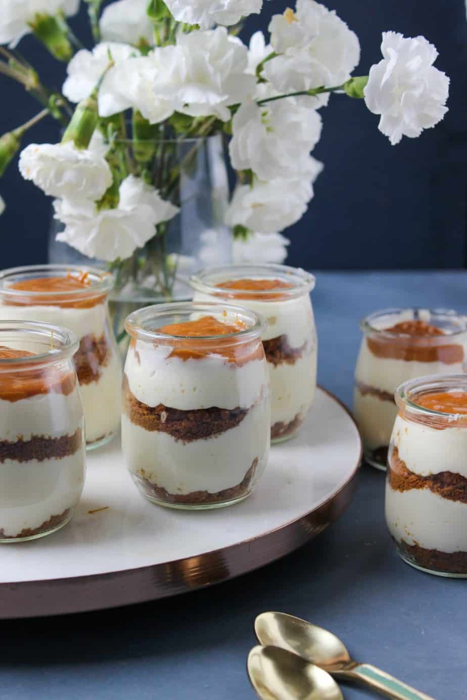 Six Cheesecake Jars filled with creamy layers and a rich brown topping are arranged on a tray in front of a vase with white flowers. Two gold spoons rest nearby on the blue surface, ready to enjoy.