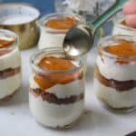 Small glass Cheesecake Jars filled with layers of cream, crumb, and caramel topping are arranged on a round marble board. A hand is about to scoop from one jar with a spoon. White flowers are visible in the background.