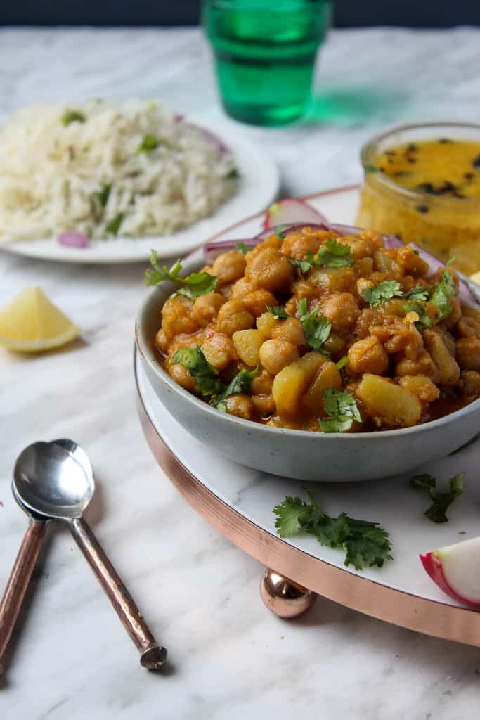 Aloo Chana with a plate of rice in the background Aloo Chole in a bowl, rice in the background, daal in a small bowl, a wedge of lemon and a glass of water.