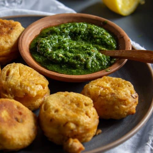 A plate of golden, crispy fritters is served with a wooden bowl of vibrant cilantro chutney. Lemon wedges are placed in the background on a gray surface.