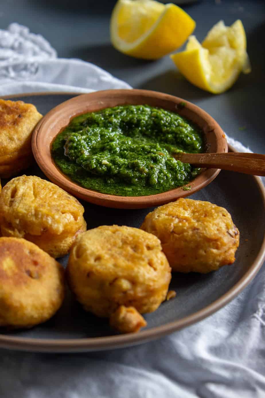 A plate of golden, crispy fritters is served with a wooden bowl of vibrant cilantro chutney. Lemon wedges are placed in the background on a gray surface.