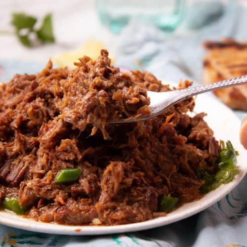 A close-up of a fork holding a portion of shredded Bhuna Gosht atop a plate, with green peppers visible underneath, set on a table with a blue cloth and glass of water in the background.