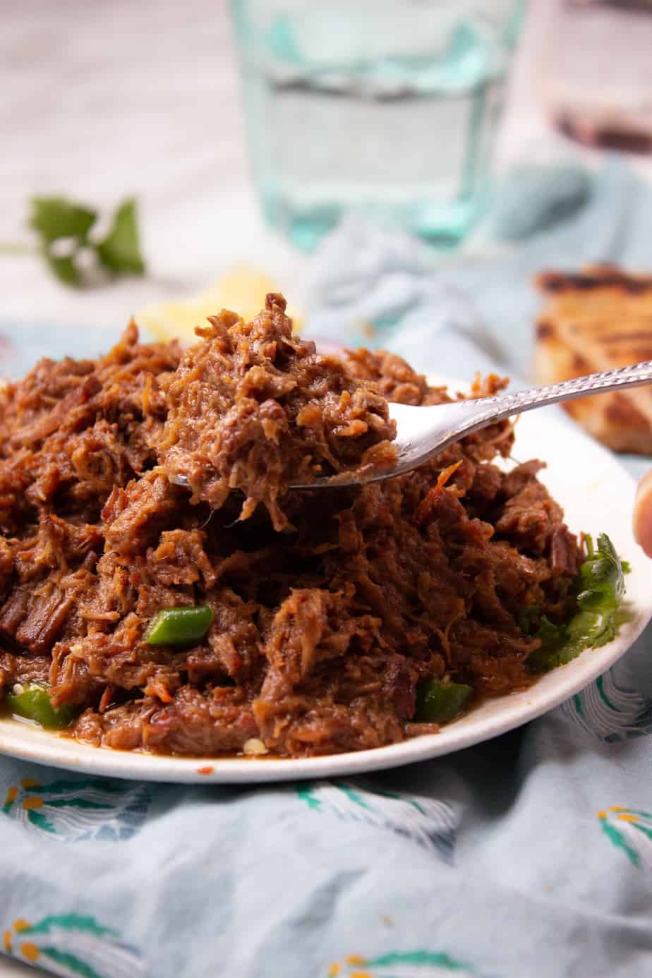 A close-up of a fork holding a portion of shredded Bhuna Gosht atop a plate, with green peppers visible underneath, set on a table with a blue cloth and glass of water in the background.