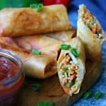 Crispy Vegetable Noodle Spring Rolls on a wooden board, some whole and some cut open to reveal noodles and veggies inside. Garnished with chopped green onions and served with a small jar of dipping sauce.