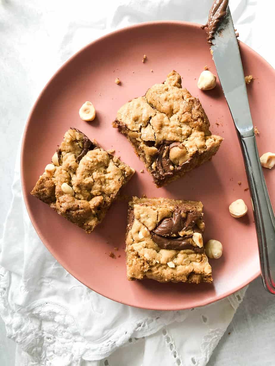 coral colored plate with 3 nutella blondies placed on it with a smattering of nuts and a knife with nutella on it