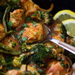 A close-up of a spicy Achari Chicken Bake with juicy chicken, broccoli florets, green chilies, and fresh cilantro, garnished with lemon slices and served in a dark pan with a metal spoon.