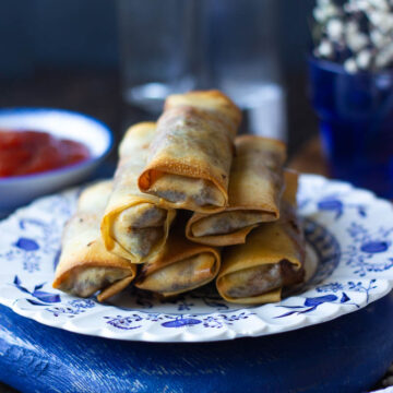 A stack of golden-brown Southwest Rolls is arranged on a decorative blue and white plate. In the background, a small bowl of red dipping sauce and blurred jars sit under soft lighting that highlights the food.