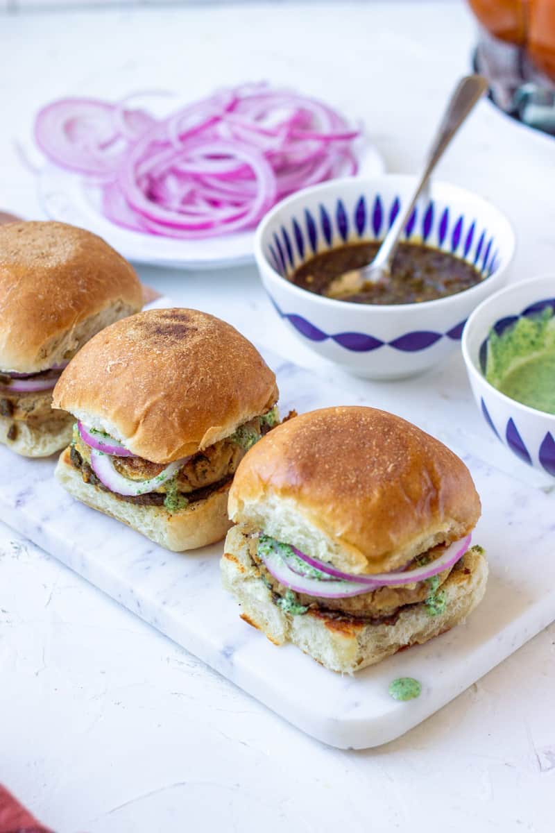 top down views of three bub kabab, partial view of one bowl of green chutney, a bowl of tamarind chutney and onions in the background