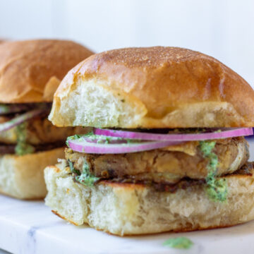 A close-up of a vegetarian Bun Kabab on a soft bun, topped with sliced red onions and a green sauce, sits on a white marble surface. Another burger is partially visible in the background.