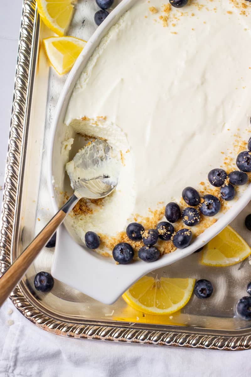 A top down view of a dish of lemon mousse with a spoonful removed and the spoon resting in its place. wedges of lemons and blueberries