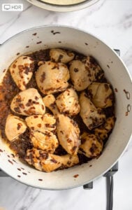 Chicken Pulao pieces simmer in a large white pot with browned onions and dark sauce, set on a stove against a marble countertop background.