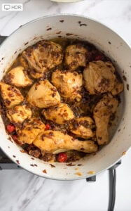 Chicken Pilau pieces cooking in a pot with browned onions, oil, and red chilies on a stovetop, photographed from above on a marble surface.