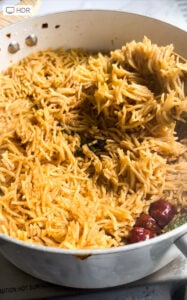 A white pan filled with aromatic Chicken Pulao, garnished with whole dried red chilies and curry leaves, sits on a stovetop. A spoon is lifting some of the spiced rice.