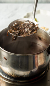 A metal ladle holds up a portion of cooked seaweed above a steaming pot of broth on a stovetop, reminiscent of the delicate preparation seen in traditional Pink tea.
