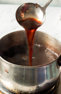 A ladle pours rich, reddish-brown sauce back into a steaming metal pot on a stovetop. The pot, darkly stained, evokes the warmth of brewing Kashmiri Chai, while the softly blurred background adds to the comforting scene.