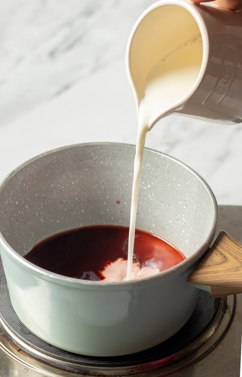 A hand pours milk from a cream-colored jug into a saucepan filled with dark red Kashmiri Chai, on a stovetop with a wooden-handled pot.