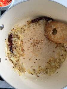 Chopped onions, cumin seeds, and dried red chilies are being sautéed in a pot for Aloo Palak. A wooden spatula stirs the lightly browned mixture as it cooks in oil.