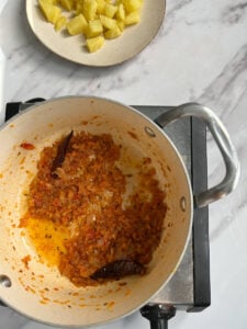 A pot of sautéed onions, tomatoes, and spices sits on a stovetop next to a plate with chunks of boiled yellow potatoes, ready for making Aloo Palak, all set on a white marble countertop.