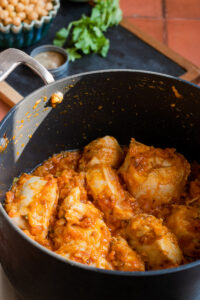 A black pot filled with Chana chicken—tender pieces simmering in a chunky orange tomato and onion sauce, with fresh cilantro, chickpeas, and a small bowl of spices in the background.