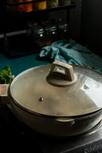 A covered saucepan with a glass lid sits on a stovetop, steam condensing inside as white chicken curry simmers. In the background, spice jars and a light blue cloth are visible on a dark kitchen counter.