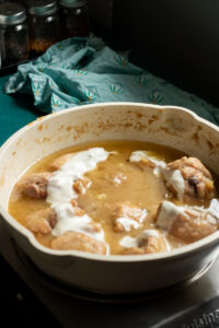 A white pot on the stove contains pieces of chicken simmering in a golden-brown sauce, reminiscent of white chicken curry, with some areas topped with a dollop of cream. Jars of spices and a green patterned cloth appear in the background.