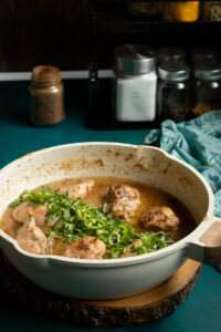 A white skillet sits on a wooden board, filled with tender white chicken curry pieces in a brown sauce, garnished with sliced green chili and fresh herbs. Spice jars and a teal cloth create a cozy background.