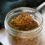 A close-up of a glass jar filled with colorful Achari Masala spice blend. A metal spoon scoops out the mixture, revealing dried herbs and crushed red pepper flakes. A dark cloth is in the background.