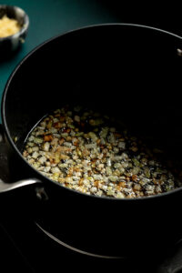 Diced onions being sautéed and caramelized in oil inside a black pot on a stovetop, as the first step in preparing authentic Pakistani Aloo Gobi, with a bowl of minced garlic blurred in the background.